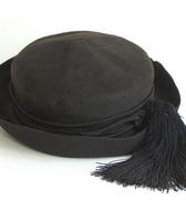 Moški klobuk iz fine črne klobučevine, z okroglim mehkim oglavjem in s kratkimi krajci brez obrobe, močno zasukanimi navzgor. Okoli oglavja je večkrat ovita črna okrogla volnena vrvica, ki se končuje z dolgima črnima cofoma / Bohinjska Češnjica / iz zbirke SEM / foto: J. Žagar. 