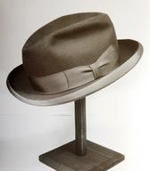 Moški klobuk iz svetlo sive klobučevine, z višjim, trikotno vtisnjenim oglavjem in s trdimi krajci, polkrožno zasukanimi navzgor (spominjajo na trdi klobuk tipa homburg). Konfekcijski izdelek tovarne Šešir, Škofja Loka. Iz družine ljubljanske modistke / Ljubljana / iz zbirke SEM / foto: J. Žagar. 
