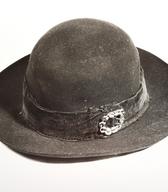 Moški klobuk z višjim okroglim oglavjem in srednjimi krajci; iz črne klobučevine, s črnim svilenim trakom okoli oglavja in okrasno kovinsko zaponko. V zadnji rabi je bil klobuk sestavina t.i. narodne noše (kostuma) / okolica Kamnika /